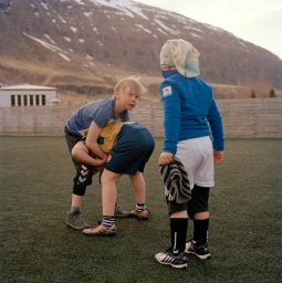 Three children playing outdoors on a green sports field with mountains in the background under natural daylight.