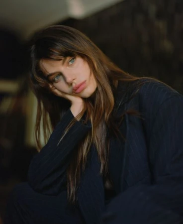 Close-up portrait of a woman with long brown hair wearing a dark blazer, resting her chin on her hand with a pensive expression.