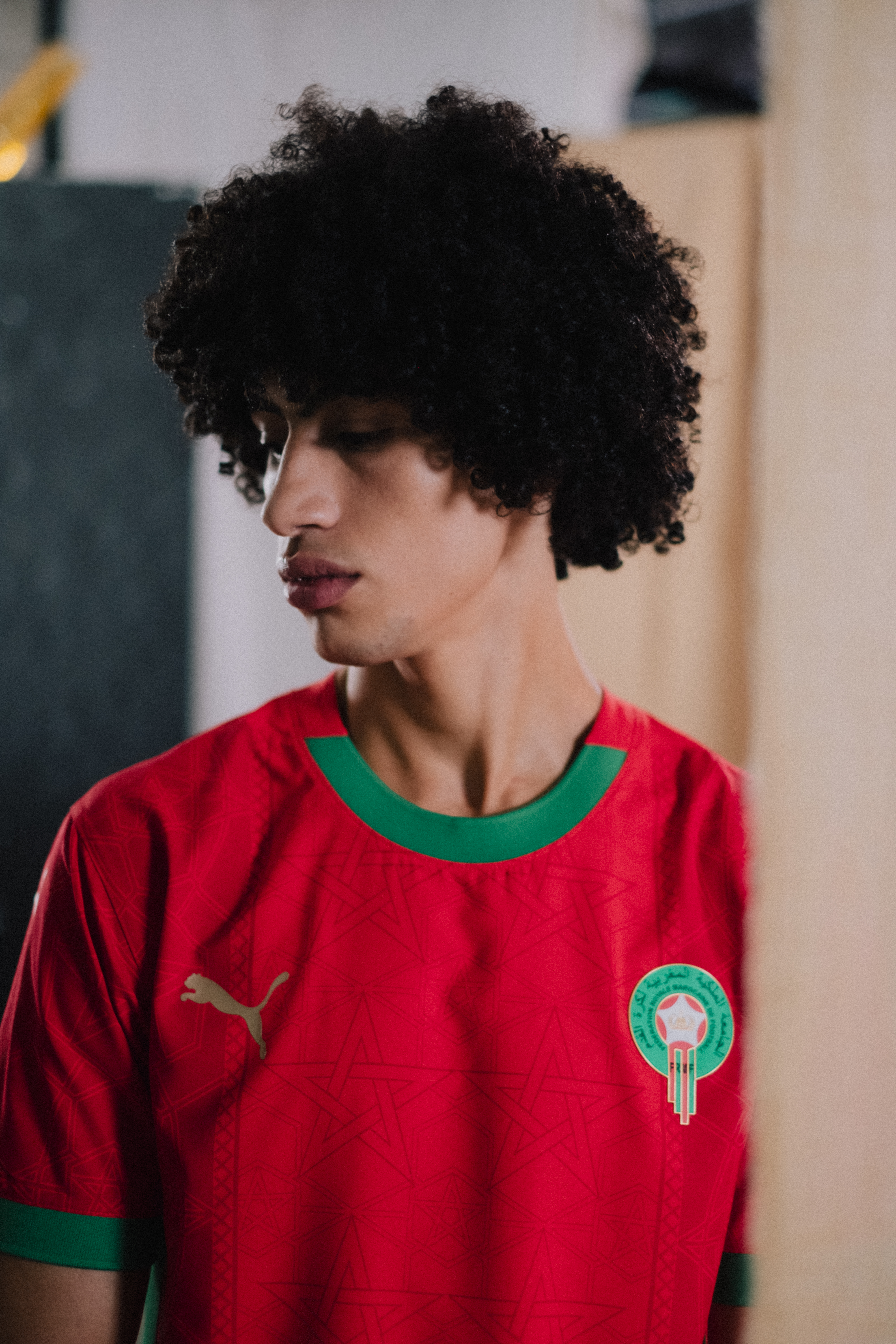 Portrait of a young adult male with curly dark hair wearing a red and green soccer jersey with the Puma logo and Morocco emblem.