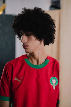 Portrait of a young adult male with curly dark hair wearing a red and green soccer jersey with the Puma logo and Morocco emblem.