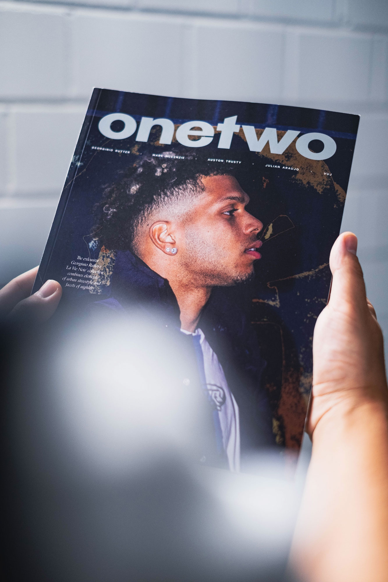 Close-up of a person holding an OneTwo magazine cover featuring a young adult male with dark hair and brown skin, wearing an earring.