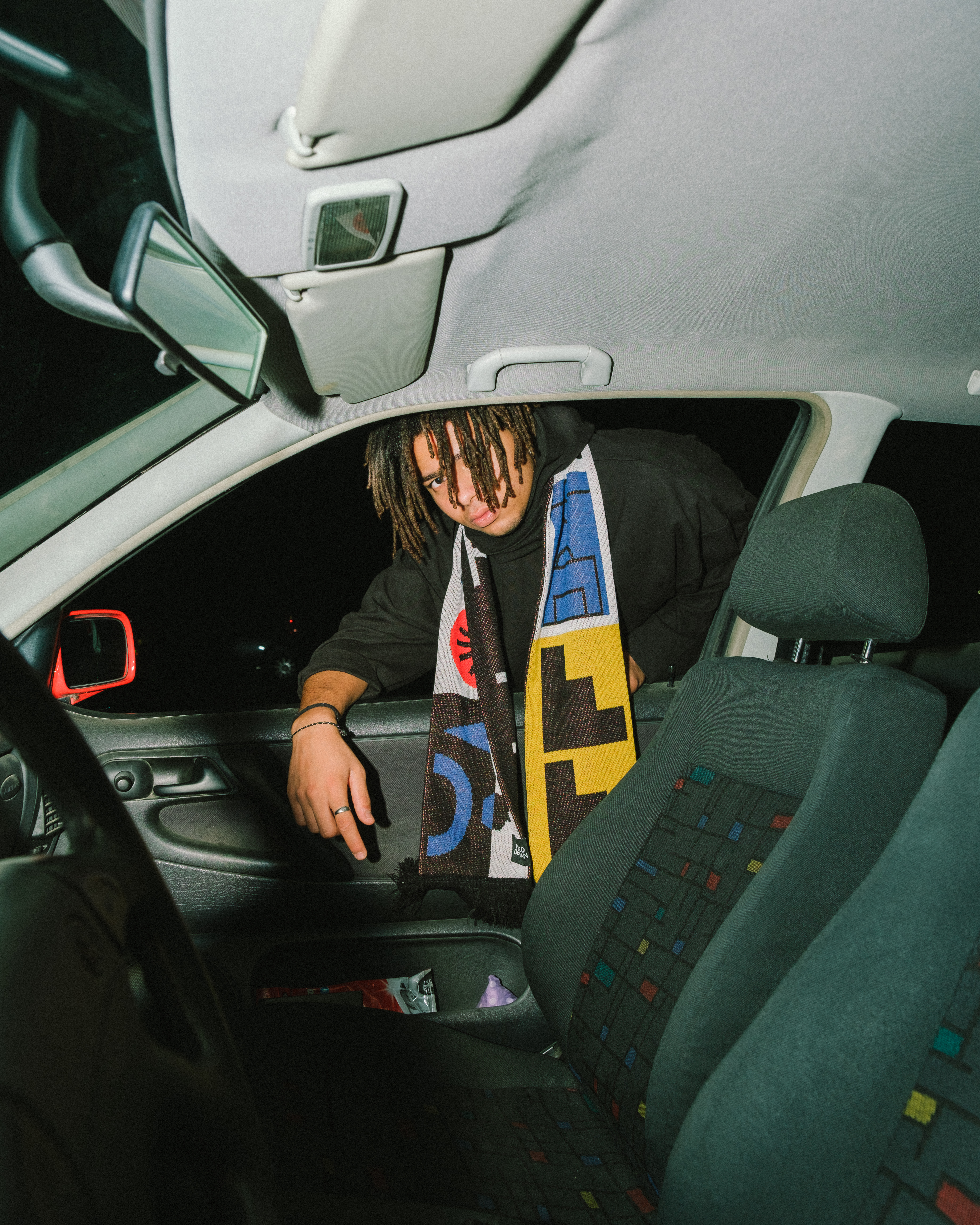 A young adult with dreadlocks leans out of a white car at night, wearing a black hoodie and a colorful patterned scarf inside a vehicle.