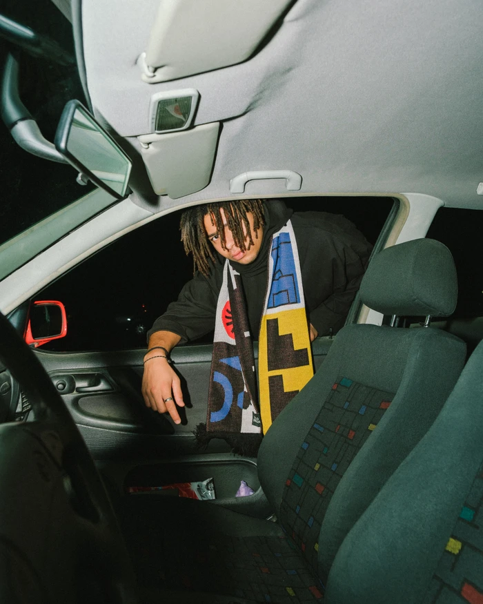 Man with dreadlocks wearing a black hoodie and graphic patterned scarf looking through an open car window at night.