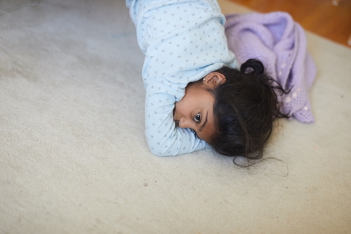 Child in pajamas lying on stomach on carpet with purple blanket next to her, looking at the camera with a soft expression.