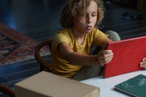Boy with blond hair and a yellow t-shirt sits at a table indoors, looking at a red tablet with a focused expression.