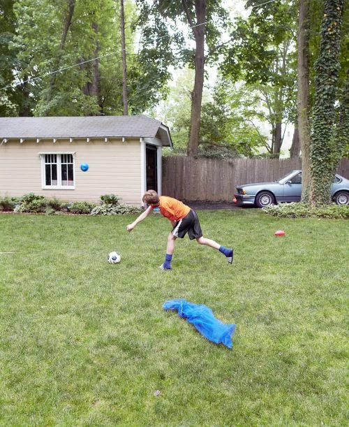 A boy in orange shirt kicks a soccer ball on a lush green lawn in a residential backyard, with a shed and a car visible.