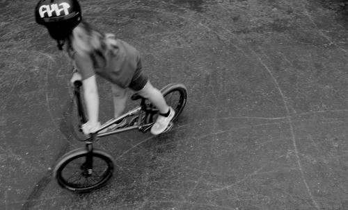 A young adult performs a trick on a BMX bike, wearing a helmet on a concrete surface, rendered in black and white, with a high angle.