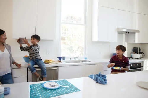Mother watching two sons in bright modern kitchen; one boy holds a tablet, the other eats at the counter. Sunny indoor lifestyle.