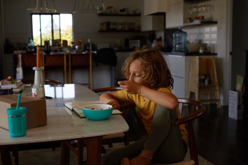 Boy with curly hair sits at a table, indoors, eating from a blue bowl with a book and beverage alongside, domestic scene.