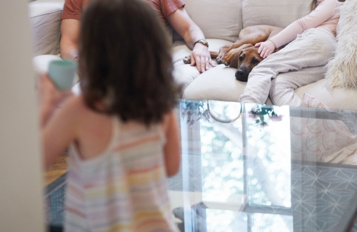 Cozy family scene indoors: adult man and child sit with a dog on a cream couch, another child stands near a glass table.
