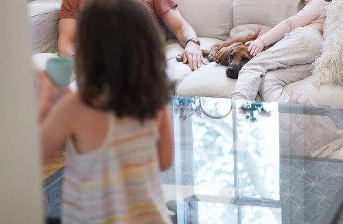 Cozy family scene indoors: adult man and child sit with a dog on a cream couch, another child stands near a glass table.