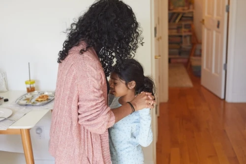 A mother with dark curly hair embraces her young daughter in a comforting hug in their well-lit home, waffles are on the table nearby.