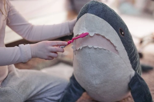 Child brushing the teeth of a plush shark toy with a pink toothbrush indoors. Grey sweatpants and long-sleeved shirt are visible.