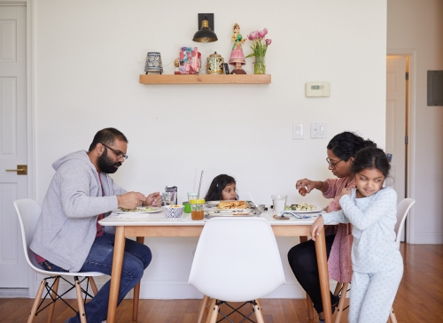 Family of four at a dining table in a bright home interior, eating and interacting. Includes food, drinks, and white chairs.
