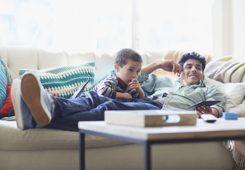 Adult male and child relax on a white sofa in a brightly lit room. Father holds a tablet and remote; child appears pensive, with a laptop nearby.