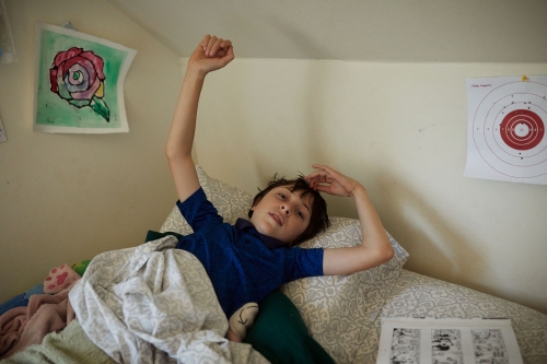 Boy in blue shirt stretches in bed surrounded by blankets and pillows with drawings of a rose and target on the wall.