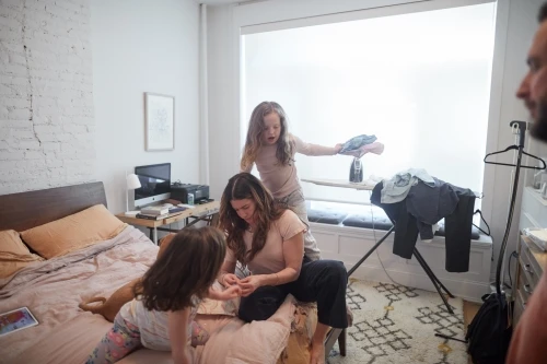 A woman sits on a bed, looking at her child's hands as another child stands nearby, with a man partially visible on the right.