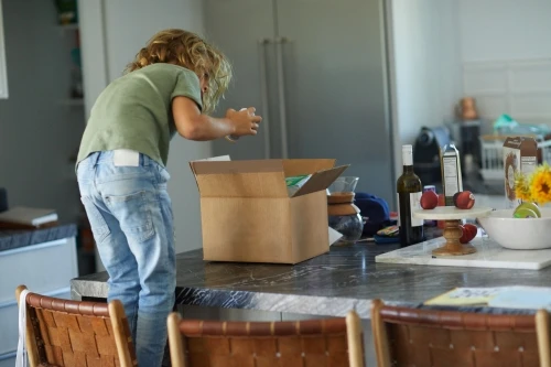 A child with blond hair standing on a chair, reaching into an open cardboard box on a kitchen counter filled with various items.