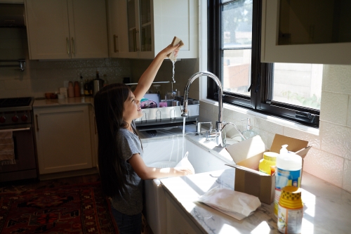 A young girl washing dishes at a kitchen sink in natural light. The white kitchen features white cabinets, a window, and various cleaning products.