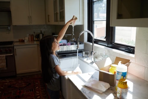 A young girl washing dishes at a kitchen sink in natural light. The white kitchen features white cabinets, a window, and various cleaning products.