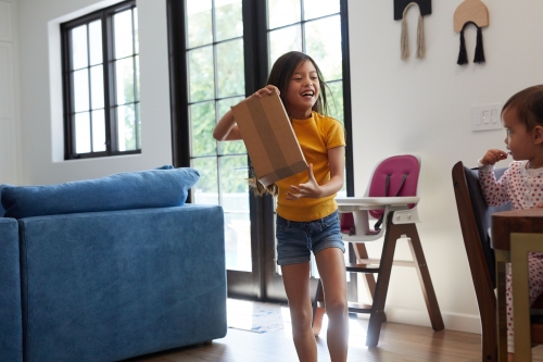 Young girl smiling and holding a box indoors, with a child seated at a table in the background, lit by daylight through a window.