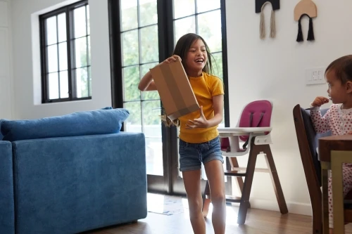 Young girl smiling and holding a box indoors, with a child seated at a table in the background, lit by daylight through a window.