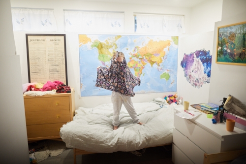 A young child stands on a bed in a bedroom, wearing a floral poncho and white pants, playing in a brightly lit indoor space.