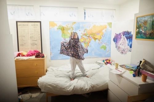 A young child stands on a bed in a bedroom, wearing a floral poncho and white pants, playing in a brightly lit indoor space.