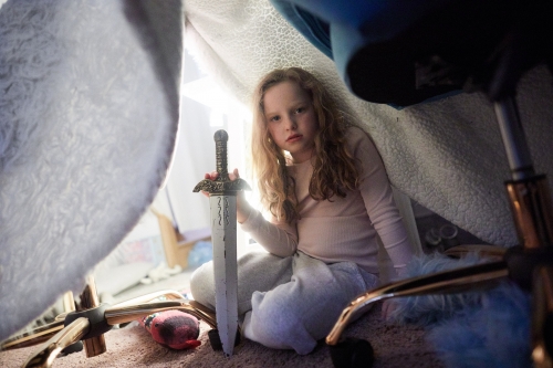 A young girl sits under blanket fort with toy sword. She's indoors, wearing pajamas, holding a large silver and gold sword, looking pensive.
