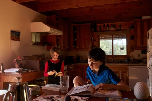 A boy reads at a wooden table while a girl reads in the kitchen, interior shot, with natural light, creating a warm, home-like ambiance.