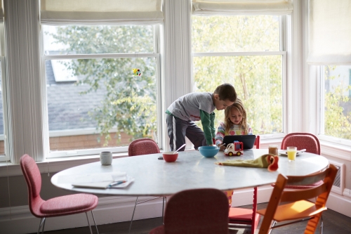 Two children, a boy and a girl, are at a round table, looking at a tablet in a brightly lit room with large windows and a view of trees.
