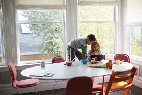 Two children, a boy and a girl, are at a round table, looking at a tablet in a brightly lit room with large windows and a view of trees.
