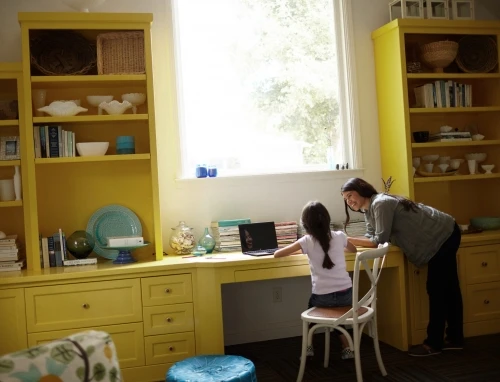 A woman and a young girl working on a laptop at a yellow desk flanked by matching yellow bookshelves in a home office.