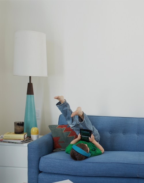 A child is lying upside down on a blue couch, playing a handheld gaming device. A lamp and white cabinet are in the background.