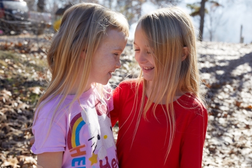 Two young girls with blonde hair embrace and smile outdoors in natural light, with a background of bare trees and foliage.