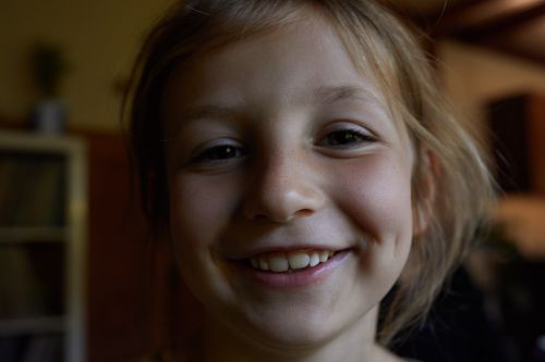 Close-up of a smiling blonde child with visible dimples, indoors with dark lighting and blurred background objects, capturing a happy portrait.
