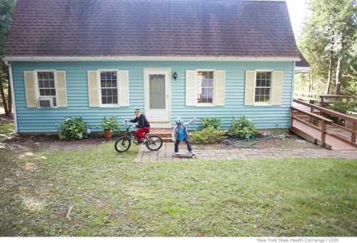 A boy rides a bike while another rides a skateboard in front of a blue house with white trim on a sunny day; outdoor activity.