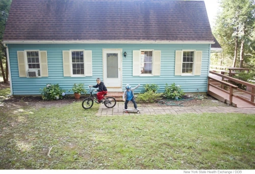 A boy rides a bike while another rides a skateboard in front of a blue house with white trim on a sunny day; outdoor activity.