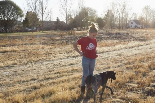 A young woman stands with her dog in a grassy field on a sunny day, wearing a red T-shirt and jeans, with trees and houses in the background.