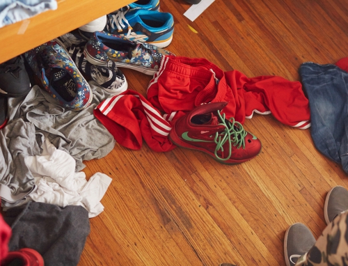Casual indoor scene of clothing and footwear scattered on a wooden floor, creating a cluttered yet familiar everyday ambiance.
