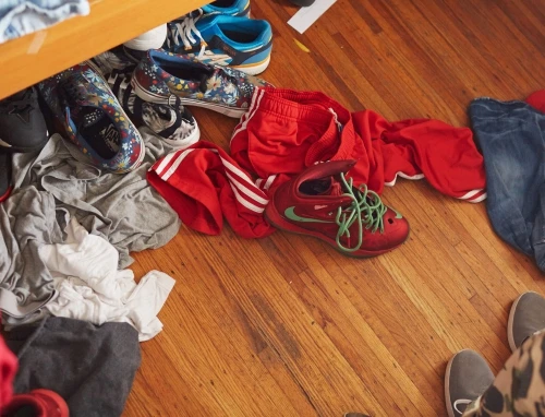 Casual indoor scene of clothing and footwear scattered on a wooden floor, creating a cluttered yet familiar everyday ambiance.