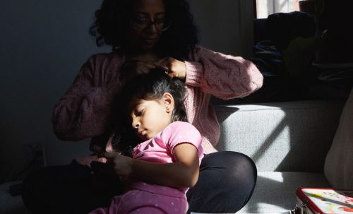 Woman braids a little girl's hair while sitting on a gray sofa in the sunlight near a window, capturing a sweet, tender moment.