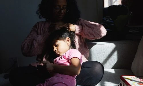 Woman braids a little girl's hair while sitting on a gray sofa in the sunlight near a window, capturing a sweet, tender moment.