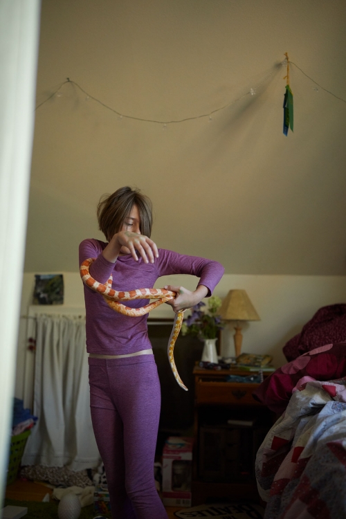 Young adult holds an orange and white corn snake indoors in a bedroom setting with a bed, lamp, and floral arrangement visible.