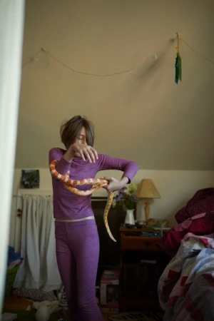Young adult holds an orange and white corn snake indoors in a bedroom setting with a bed, lamp, and floral arrangement visible.