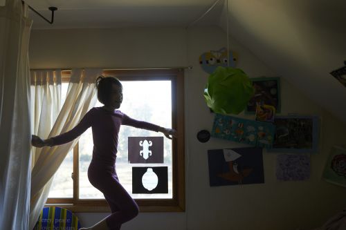A young girl in pajamas is silhouetted by a window with curtains in a bedroom setting with art and a green paper lantern.