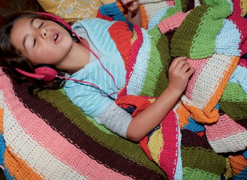 A child with pink headphones lying on a colorful striped knitted blanket. The child's eyes are closed and mouth is slightly open.