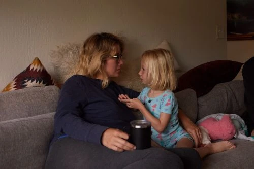 A woman and child sitting on a couch with pillows. The woman is holding a dark colored mug and the child is reaching out to the woman.