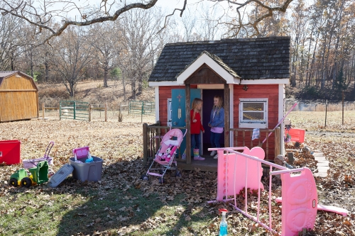 Two young girls standing on the porch of a red playhouse in a grassy backyard with scattered toys on a sunny day outdoors.