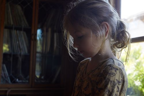 Profile portrait of a young child with a ponytail wearing a patterned top indoors near a wooden cabinet.
